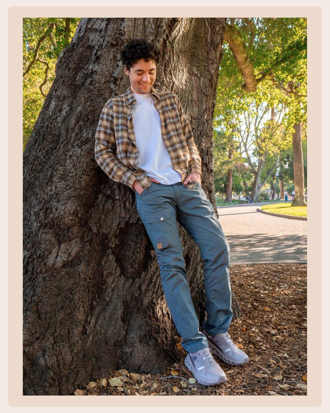 Man wearing gray On sneakers with blue-green pants and a plaid overshirt, leaning against a large tree in a sunny park.