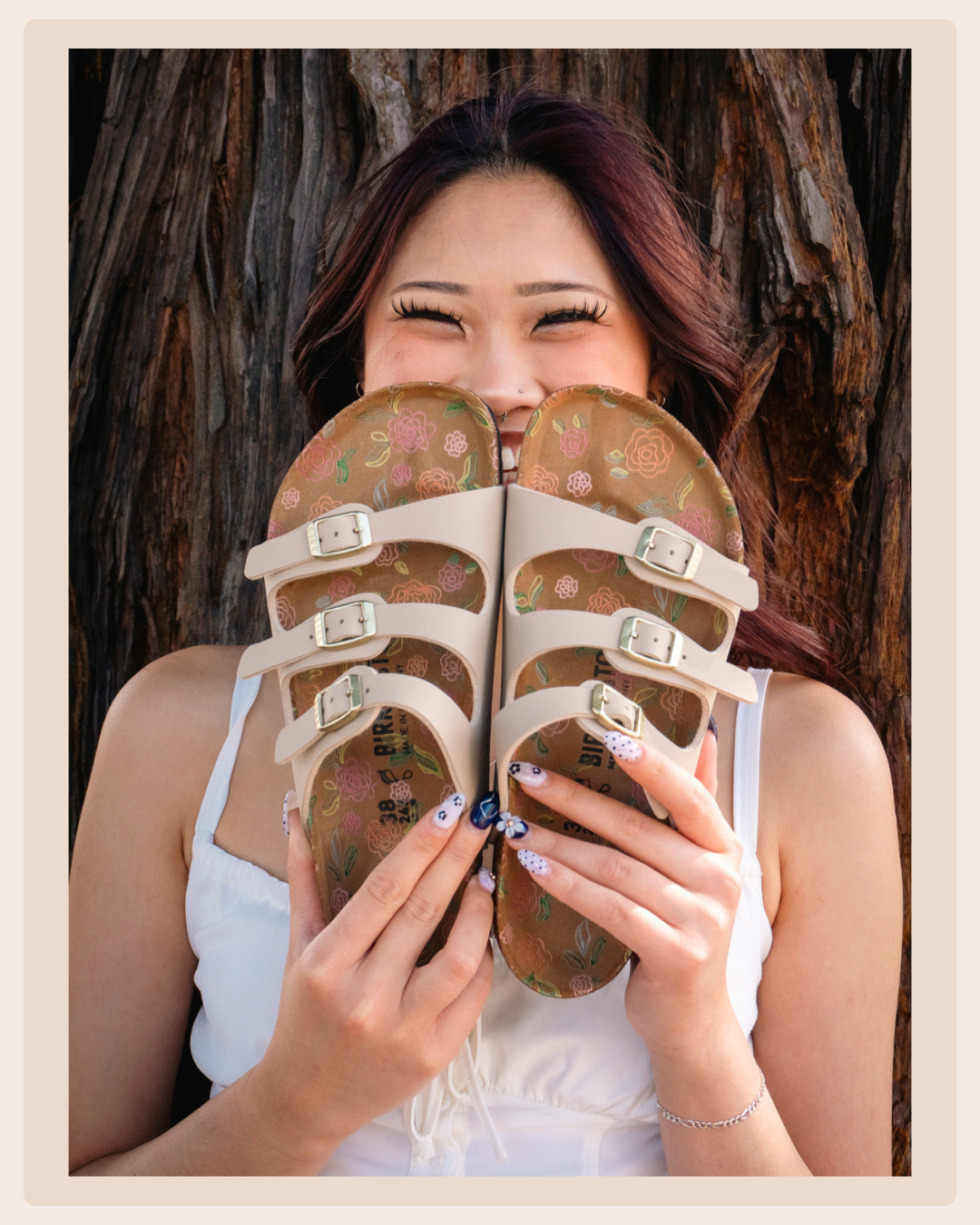 Woman holding a pair of beige Birkenstock sandals with floral footbeds in front of her face while smiling outdoors.