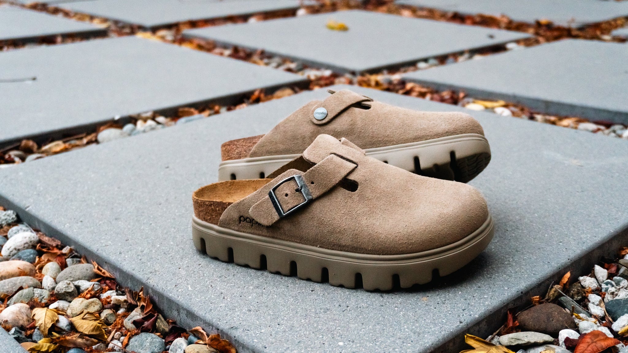 A pair of taupe-brown clog-style shoes with a single strap and silver buckle, resting on gray square patio stones outdoors with pebbles and fallen leaves around them.